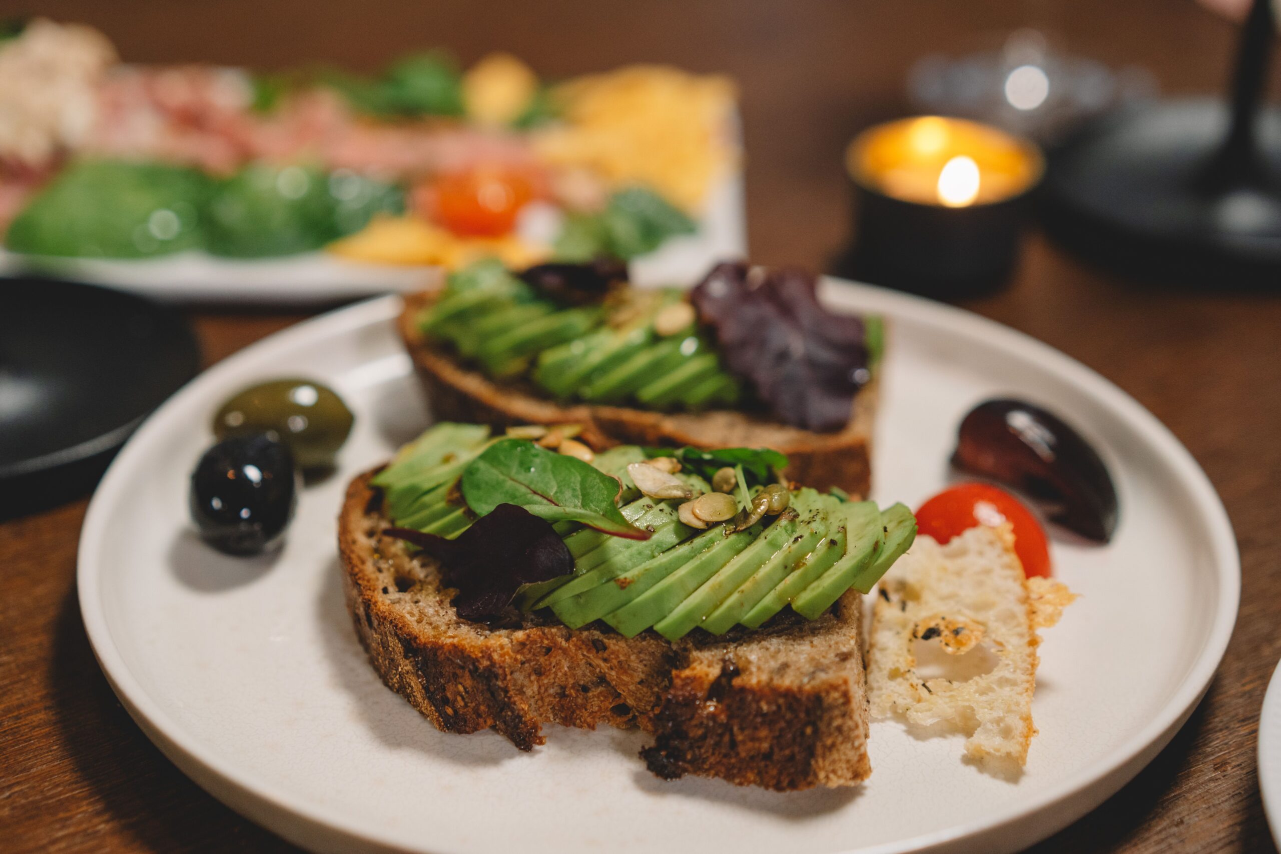 Delicious avocado toast topped with fresh greens and seeds, served with olives on a white plate.